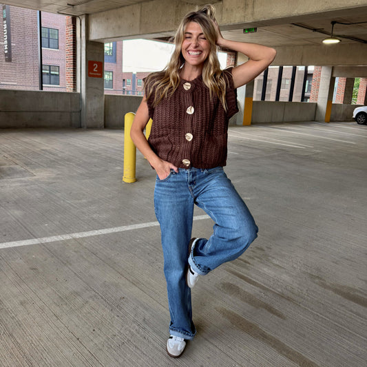 Woman posing in a parking garage wearing a brown button-up shirt and blue jeans.